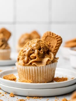 A cupcake topped with a generous swirl of Biscoff buttercream, drizzled with cookie butter, sprinkled with Biscoff cookie crumbs, and garnished with a whole Lotus Biscoff cookie, served on a white plate