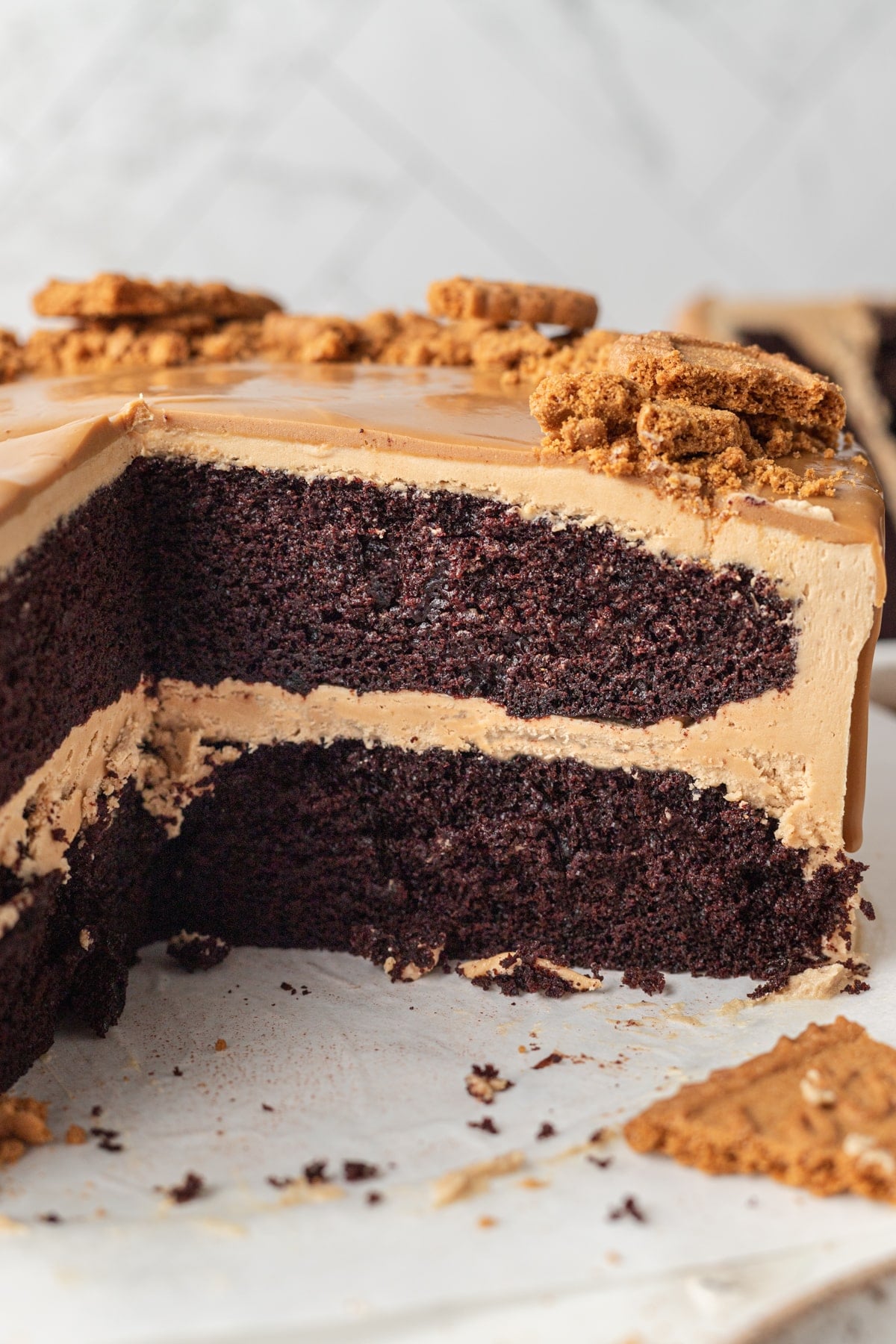 A two-layer chocolate Biscoff cake sliced open on parchment paper, revealing dark chocolate cake layers filled and frosted with Biscoff buttercream, topped with a cookie butter drizzle and crushed Biscoff cookies