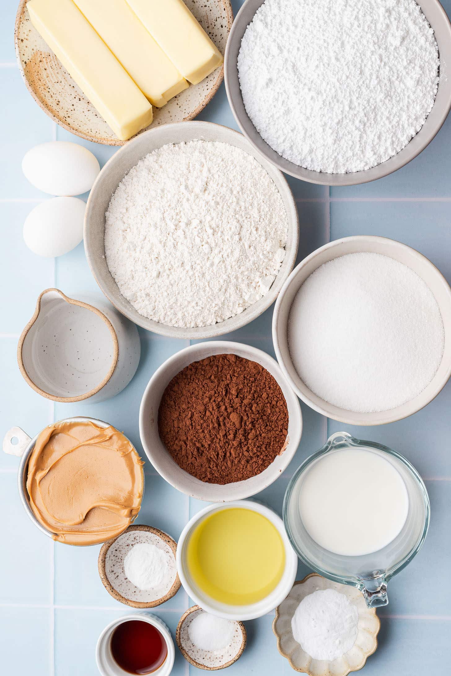 Overhead view of chocolate peanut butter cake ingredients arranged on a light blue tile surface, including butter, flour, powdered sugar, granulated sugar, cocoa powder, eggs, milk, peanut butter, oil, vanilla extract, and leavening agents