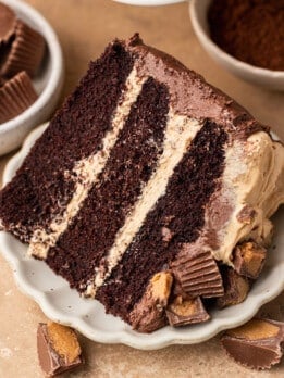 A generous slice of three-layer chocolate peanut butter cake on a white scalloped plate, showing thick peanut butter buttercream between each chocolate cake layer, surrounded by chopped Reese's peanut butter cups