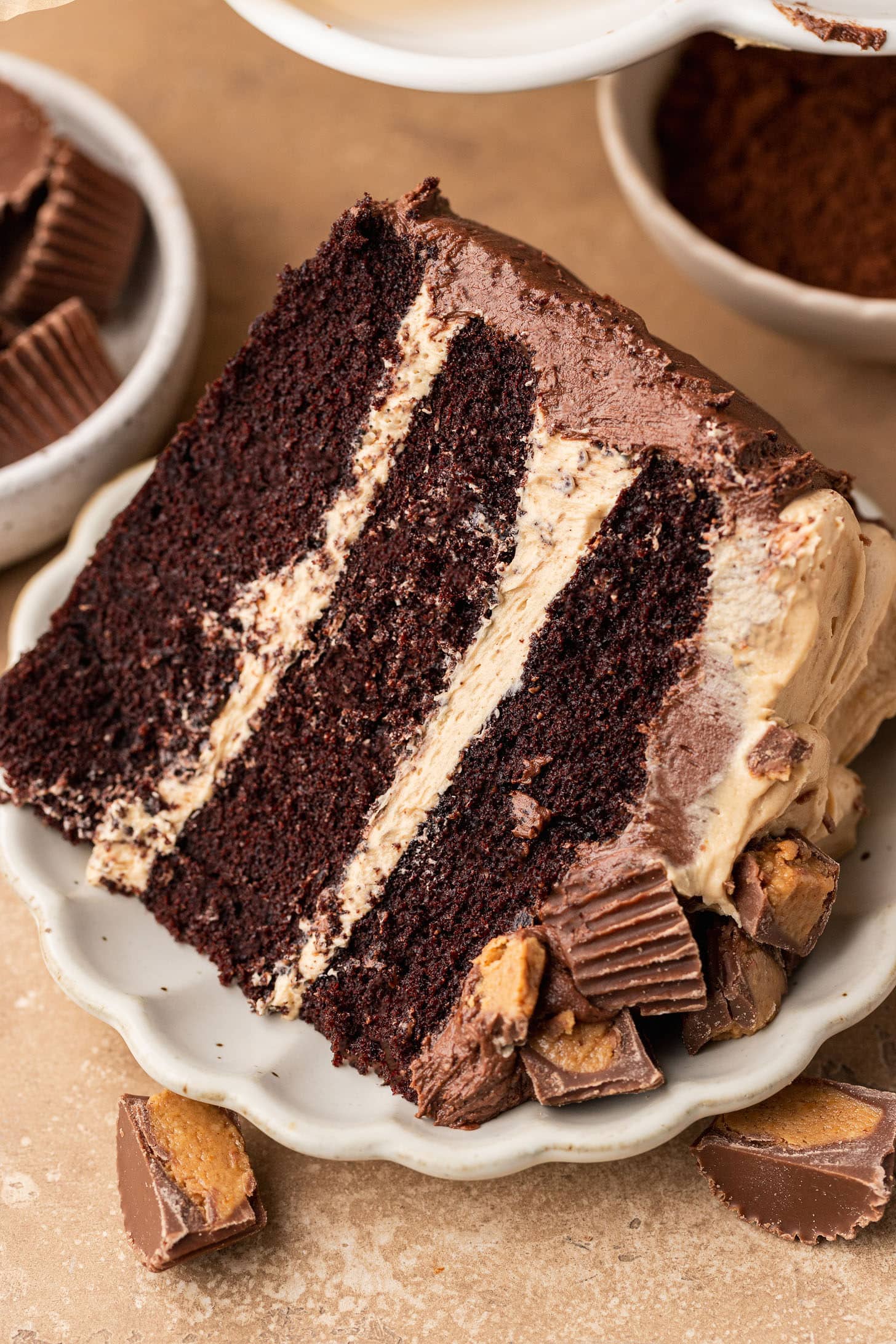 A generous slice of three-layer chocolate peanut butter cake on a white scalloped plate, showing thick peanut butter buttercream between each chocolate cake layer, surrounded by chopped Reese's peanut butter cups