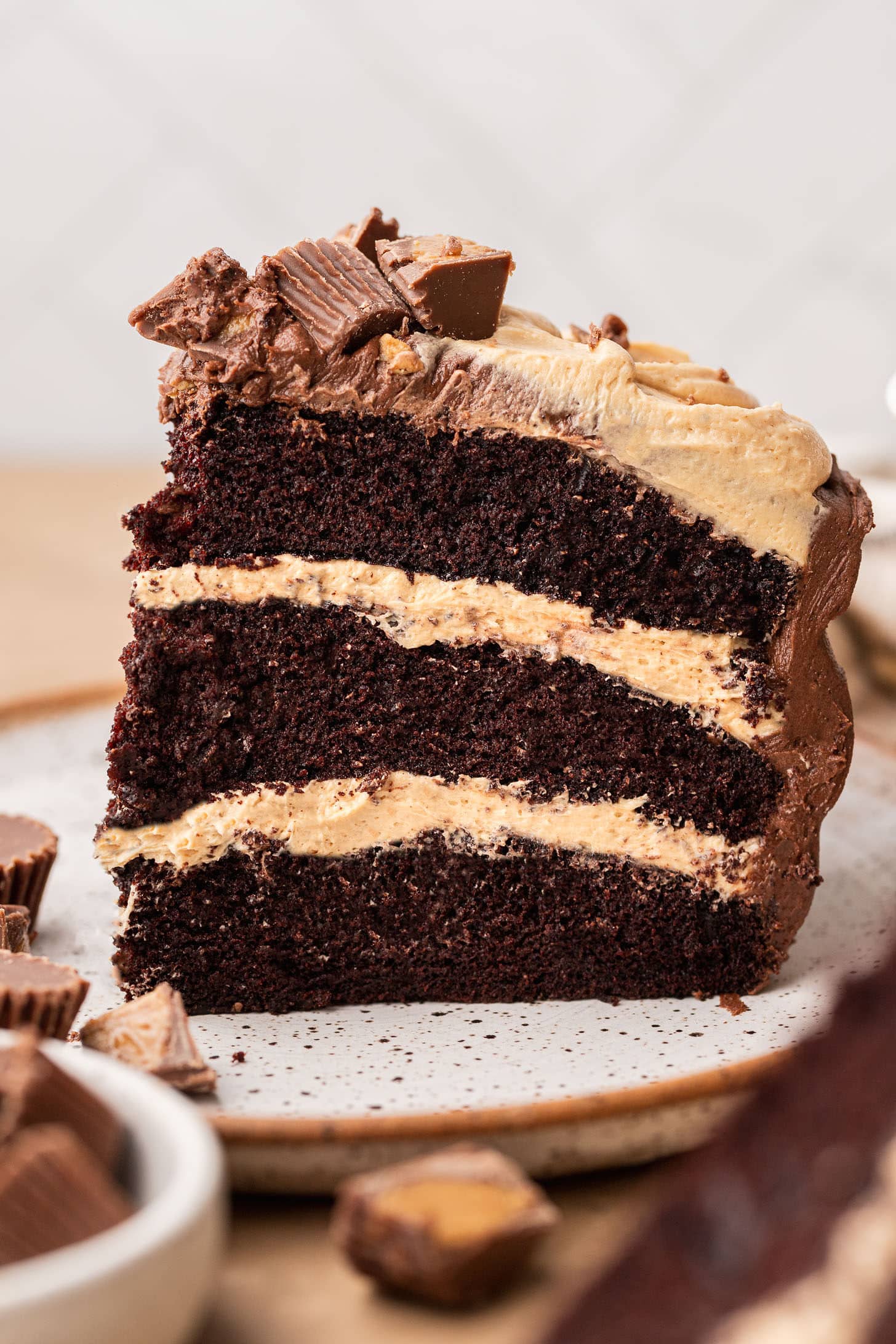 A tall slice of three-layer chocolate peanut butter cake on a speckled plate, showing dark chocolate cake layers separated by thick peanut butter buttercream, topped with piped frosting and chopped Reese's cups, with more candy pieces scattered around