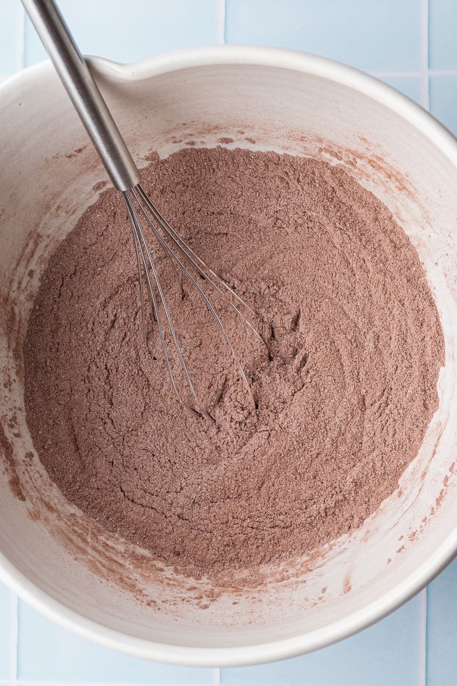 Overhead view of dry chocolate cake ingredients whisked together in a large white ceramic bowl, showing a light brown cocoa and flour mixture with a stainless steel whisk