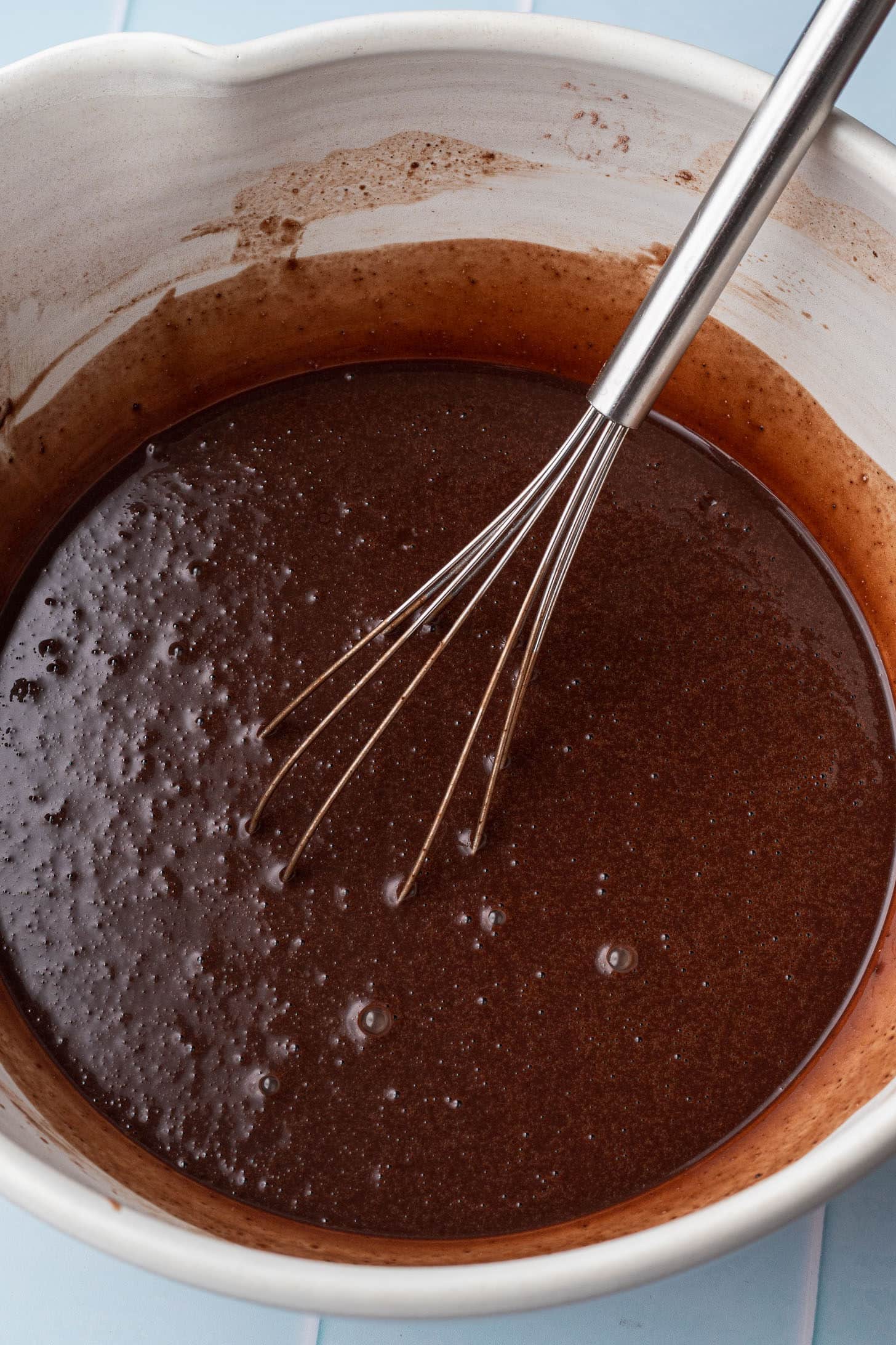 Overhead view of smooth, thin chocolate cake batter in a large white ceramic bowl being mixed with a whisk