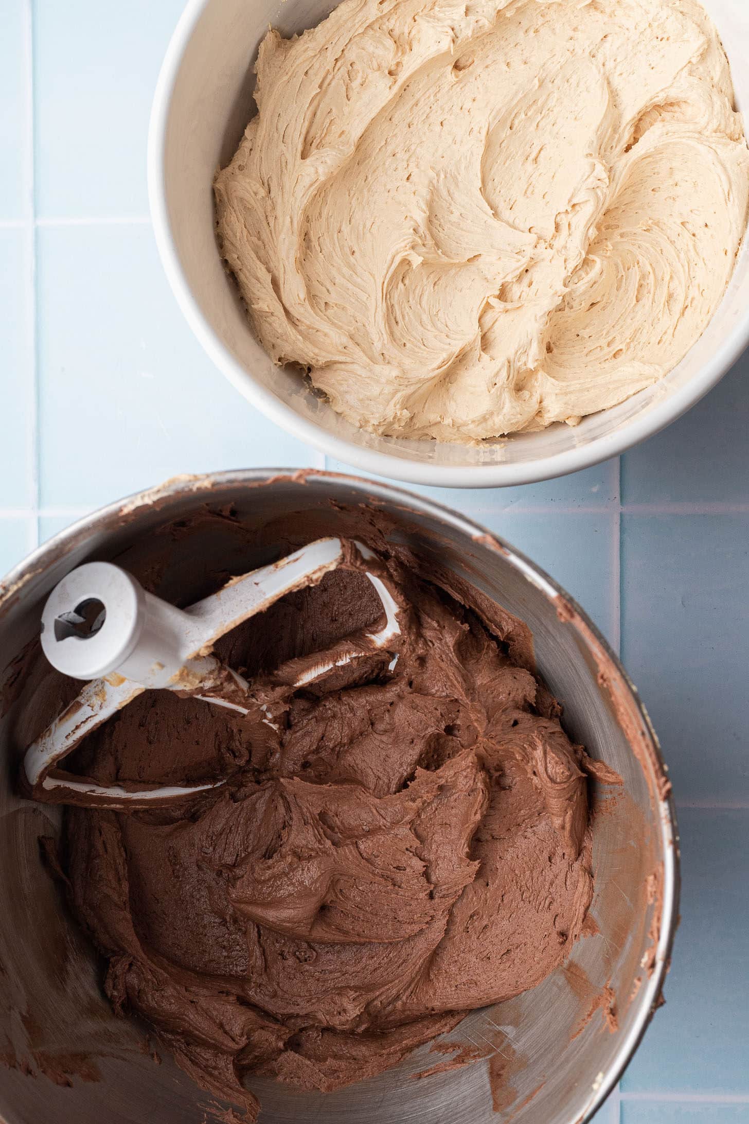 Overhead view of two bowls of frosting side by side on a light blue tile surface &mdash; a bowl of fluffy peanut butter buttercream and a stand mixer bowl of chocolate buttercream with a paddle attachment