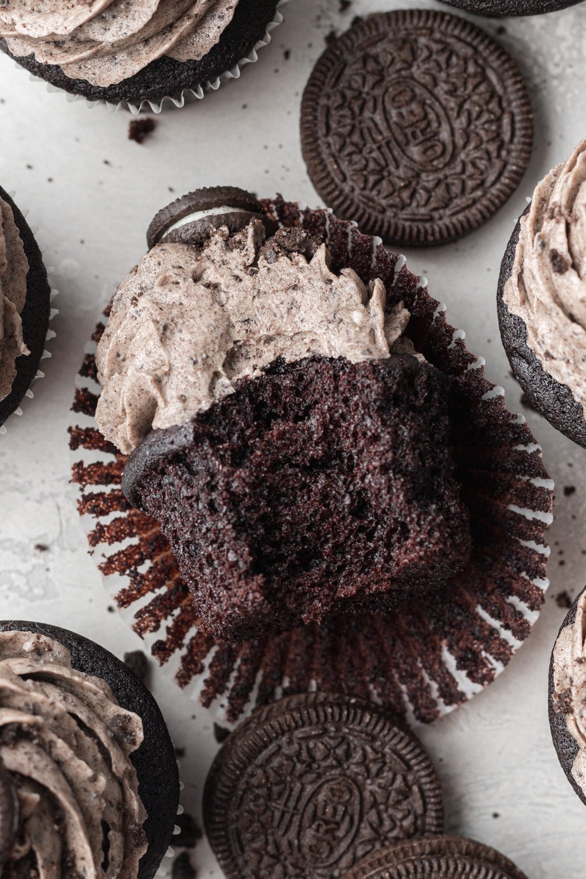 A chocolate cupcake broken open to reveal a rich, dark crumb, topped with swirled Oreo buttercream and a mini Oreo cookie, surrounded by whole Oreo cookies