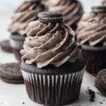 Chocolate cupcakes topped with tall swirls of Oreo buttercream and a mini Oreo cookie on top, surrounded by whole Oreo cookies and cookie crumbs on a light surface