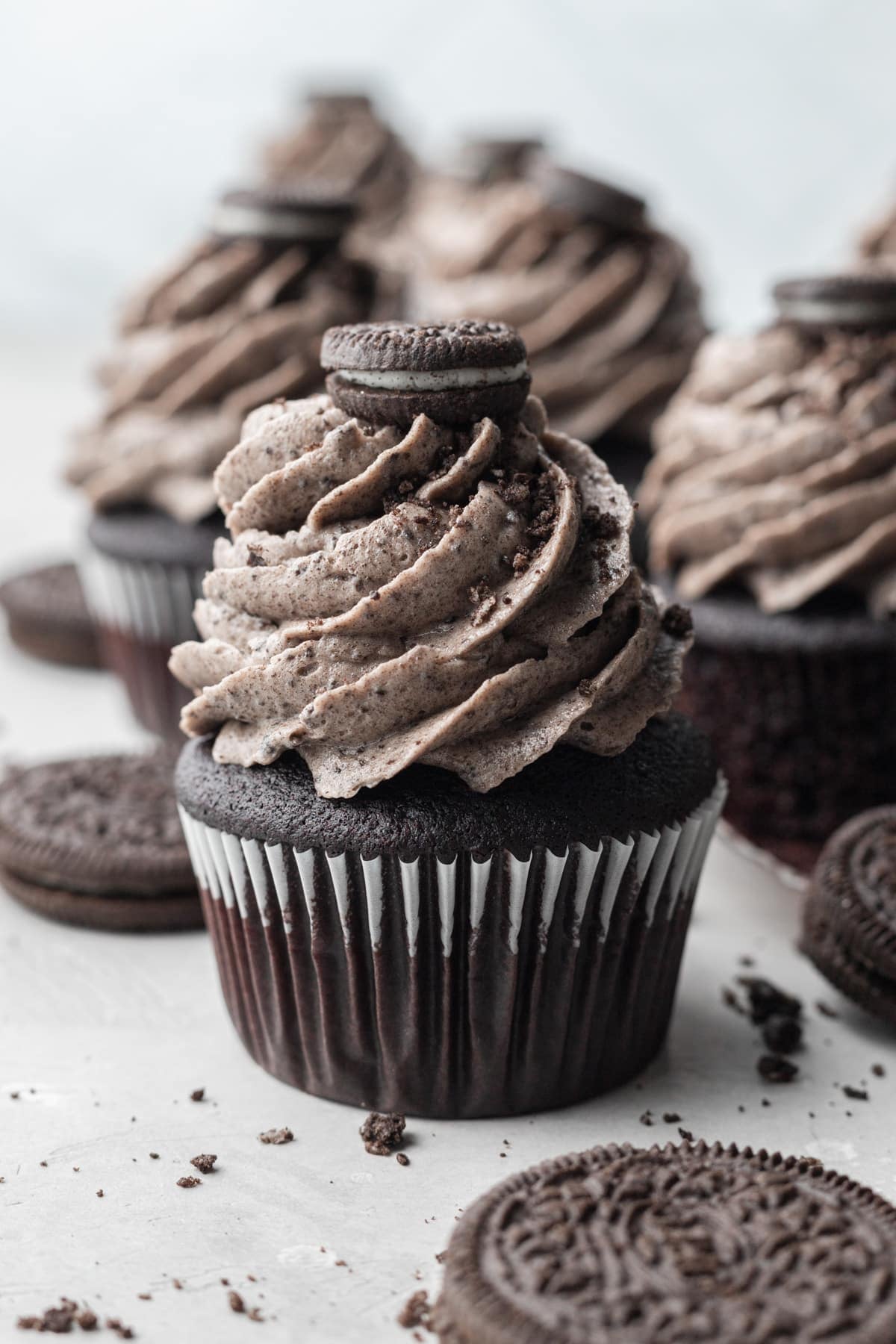 Chocolate cupcakes topped with tall swirls of Oreo buttercream and a mini Oreo cookie on top, surrounded by whole Oreo cookies and cookie crumbs on a light surface