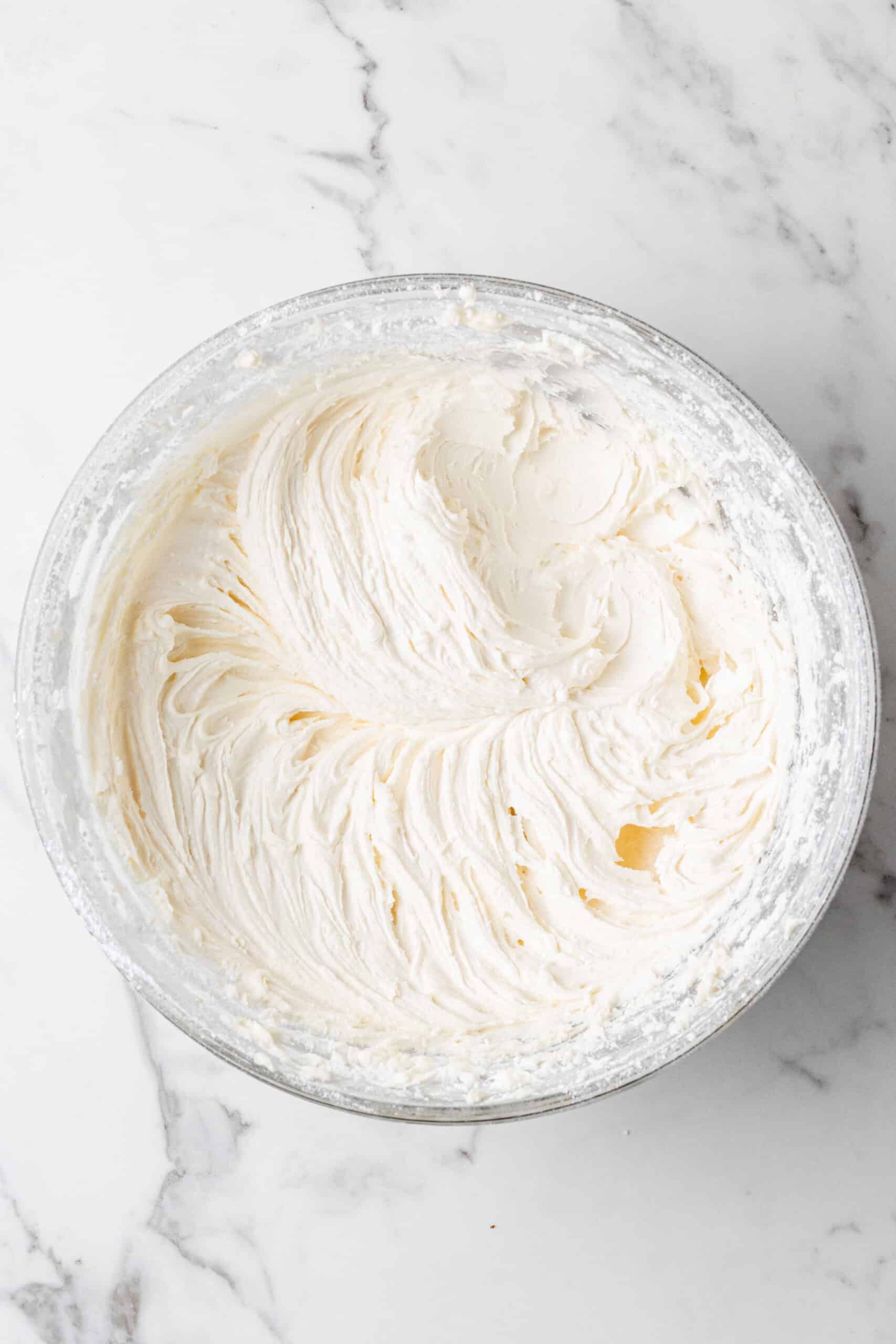 Fluffy white vanilla buttercream with powdered sugar fully incorporated, whipped to a light and airy consistency in a glass bowl on a white marble surface