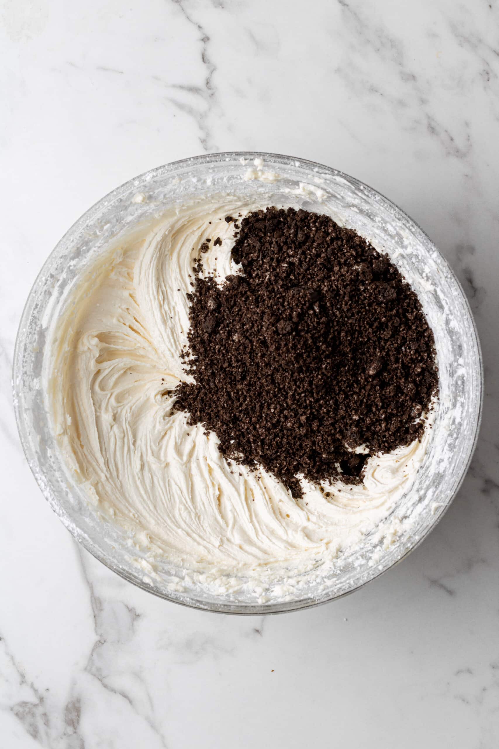 Crushed Oreo cookie crumbs added to white vanilla buttercream in a glass mixing bowl, ready to be folded in