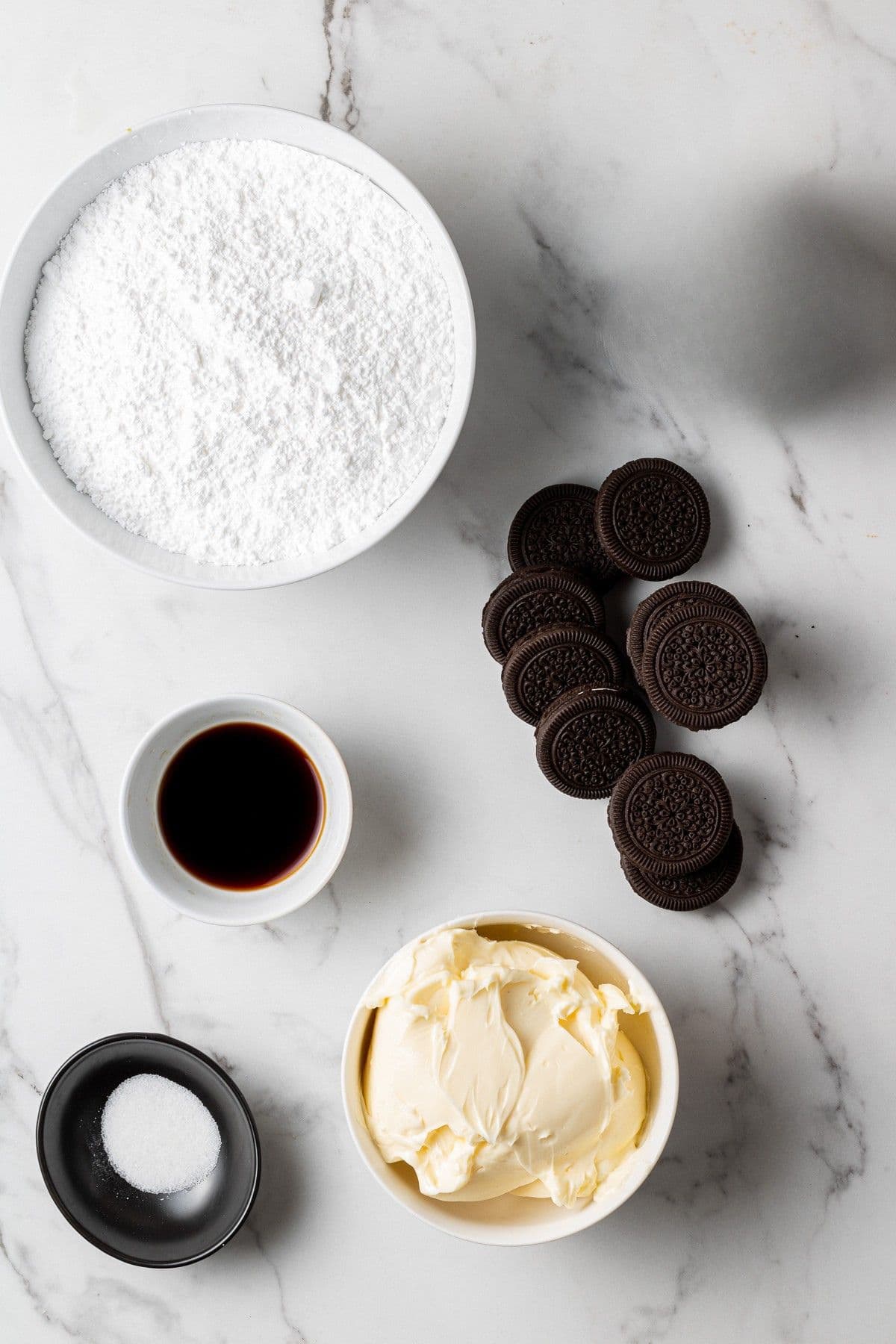 Flat lay of Oreo buttercream ingredients including powdered sugar, Oreo cookies, softened butter, vanilla extract, and salt on a white marble surface