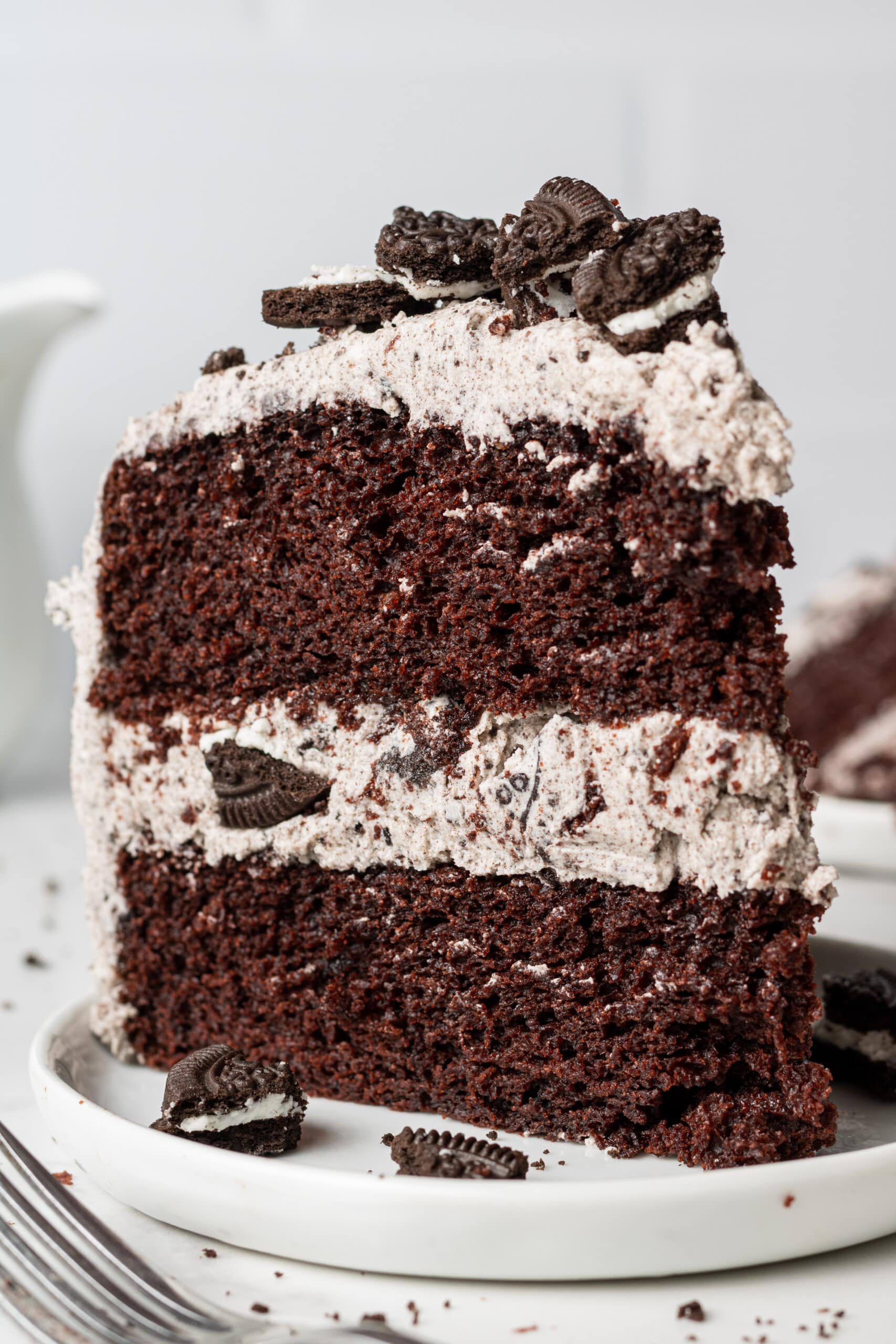 A tall slice of layered chocolate cake filled and frosted with Oreo buttercream, topped with crushed Oreo pieces and mini Oreo cookies, on a white plate