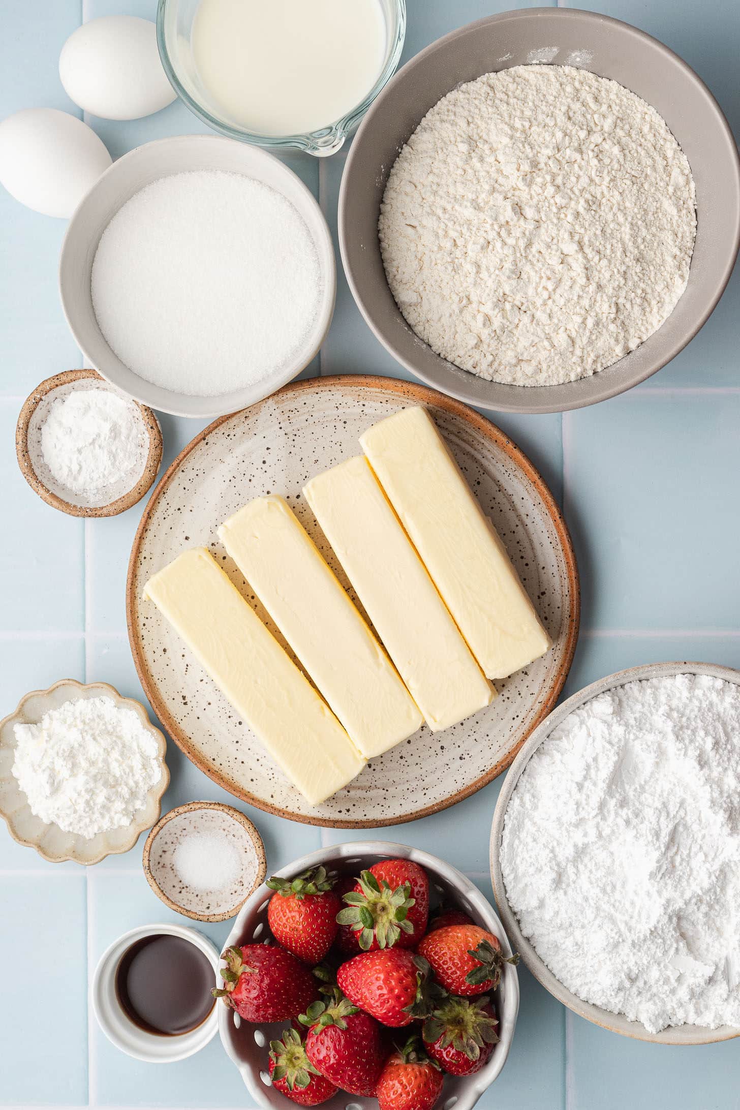 Flat lay of strawberry cupcake ingredients including butter, eggs, flour, sugar, fresh strawberries, powdered sugar, cornstarch, salt, vanilla extract, and milk on a light blue tile surface