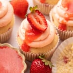 Overhead close-up of strawberry cupcakes with swirled pink buttercream and fresh strawberry slices, surrounded by whole strawberries and a small dish of strawberry puree