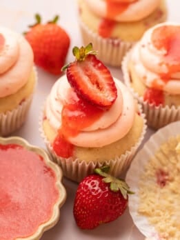 Overhead close-up of strawberry cupcakes with swirled pink buttercream and fresh strawberry slices, surrounded by whole strawberries and a small dish of strawberry puree