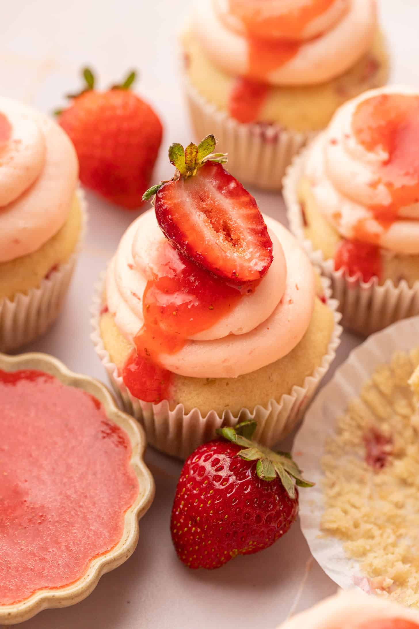 Overhead close-up of strawberry cupcakes with swirled pink buttercream and fresh strawberry slices, surrounded by whole strawberries and a small dish of strawberry puree