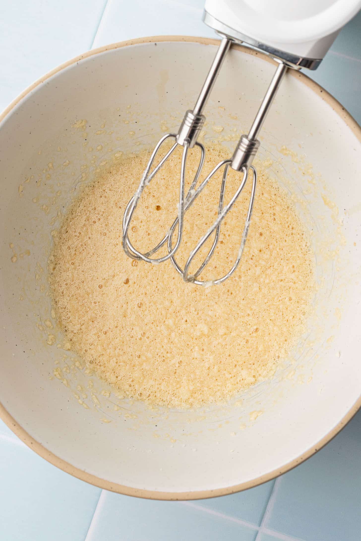 Hand mixer beating butter, eggs, and sugar together in a cream-colored ceramic mixing bowl until light and foamy