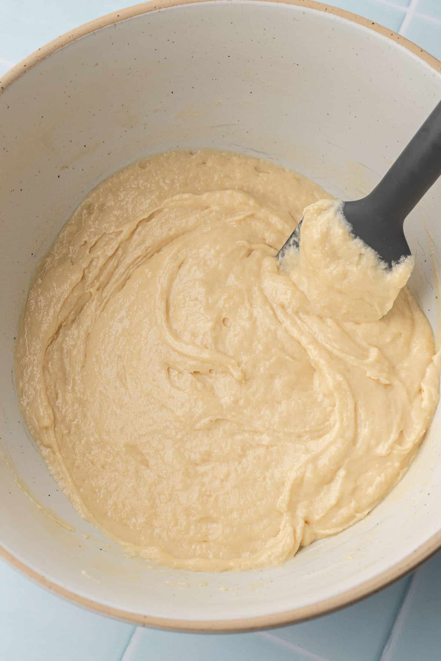 Thick vanilla cupcake batter in a cream-colored ceramic bowl being folded with a black spatula