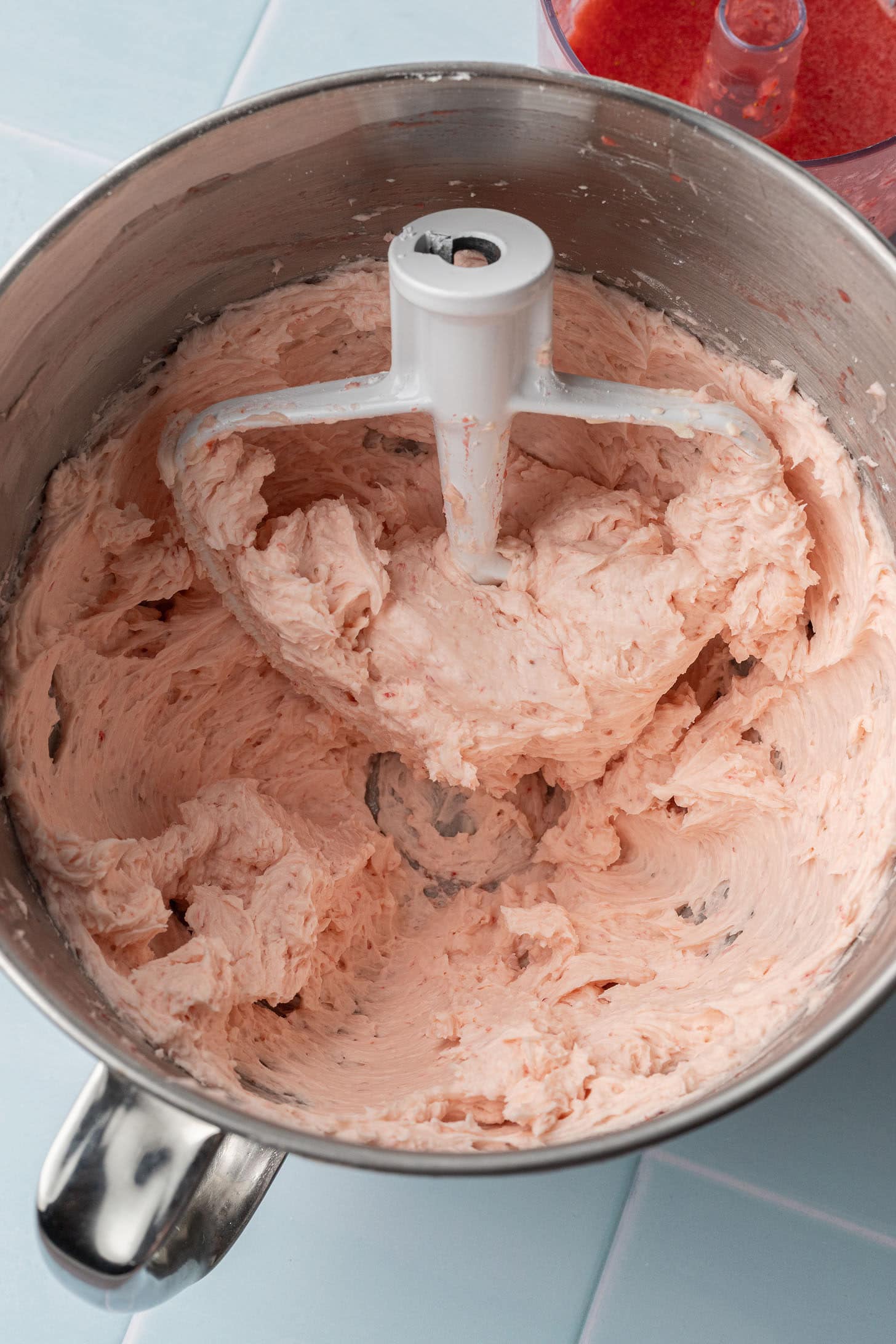 Fluffy pink strawberry buttercream being mixed in a stand mixer bowl with fresh strawberry puree visible in the background