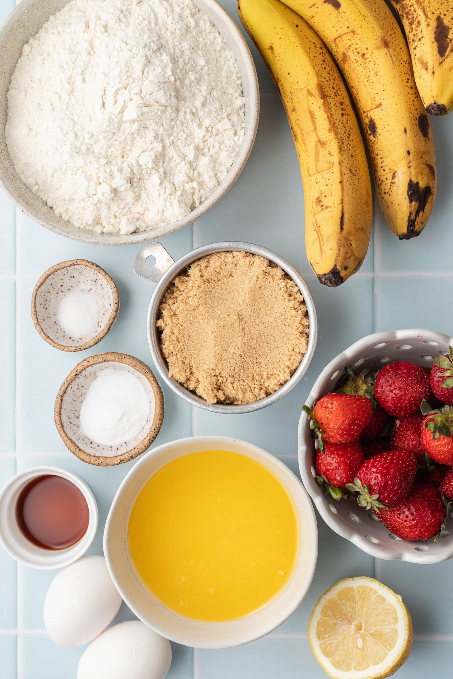 Flat lay of strawberry banana muffin ingredients including flour, overripe bananas, fresh strawberries, brown sugar, melted butter, eggs, baking soda, salt, vanilla extract, and a halved lemon on a light blue tile surface