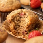 A strawberry banana muffin broken open in its wrapper to reveal a soft, moist crumb packed with roasted strawberry pieces, surrounded by more muffins and fresh strawberries