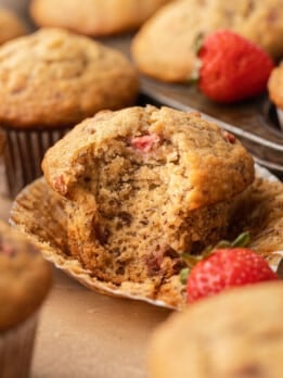 A strawberry banana muffin broken open in its wrapper to reveal a soft, moist crumb packed with roasted strawberry pieces, surrounded by more muffins and fresh strawberries