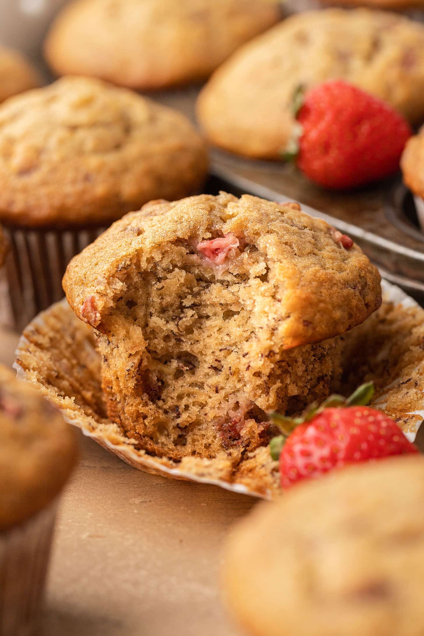 A strawberry banana muffin broken open in its wrapper to reveal a soft, moist crumb packed with roasted strawberry pieces, surrounded by more muffins and fresh strawberries