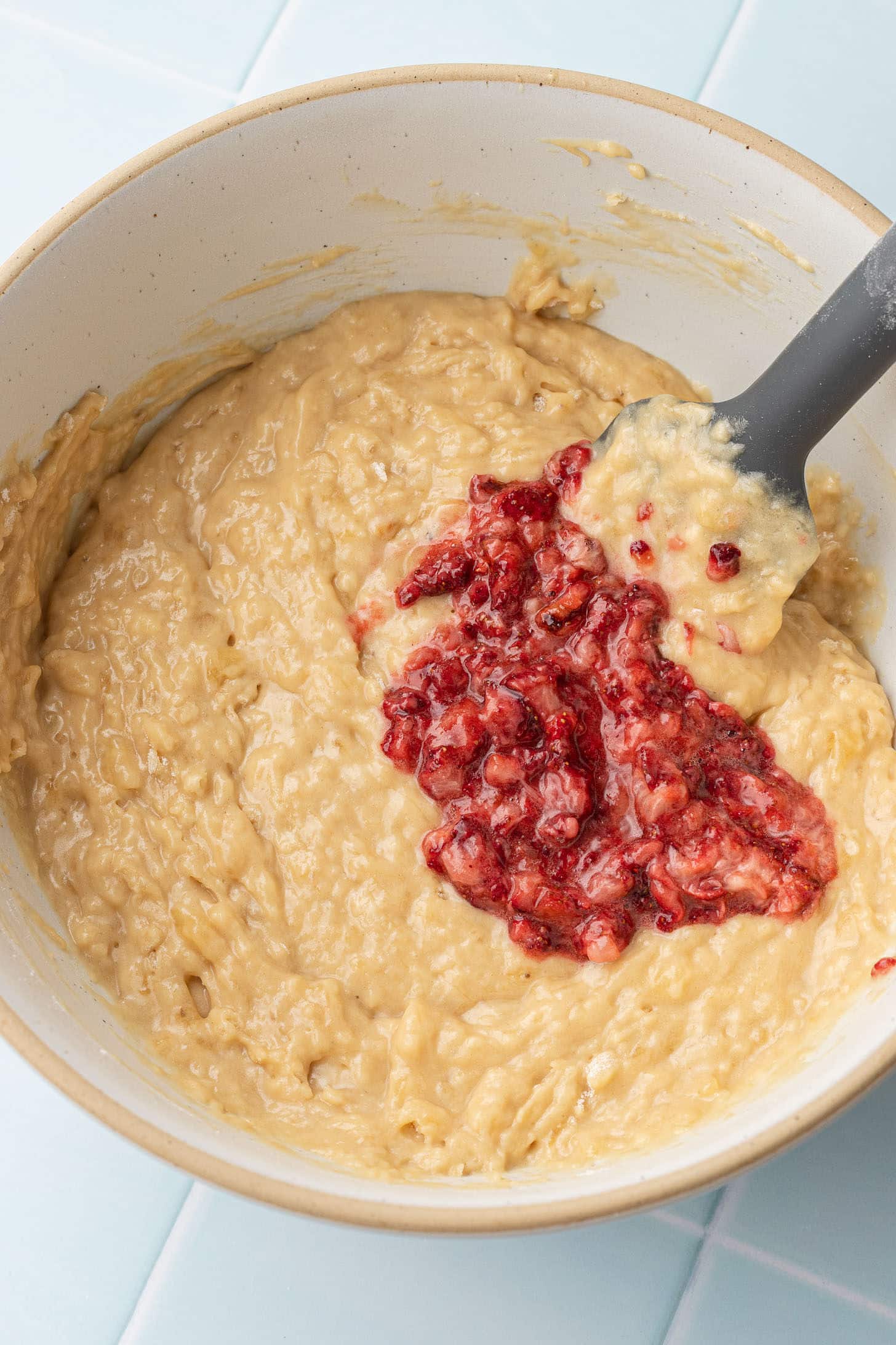Roasted strawberries being folded into banana muffin batter in a ceramic bowl with a black spatula