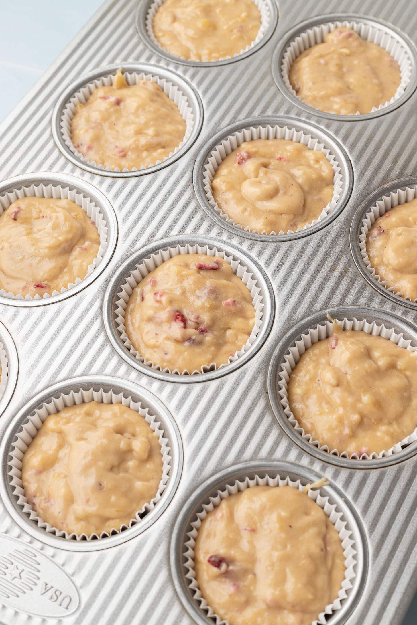 Strawberry banana muffin batter divided into a silver muffin tin lined with white parchment liners, ready to bake