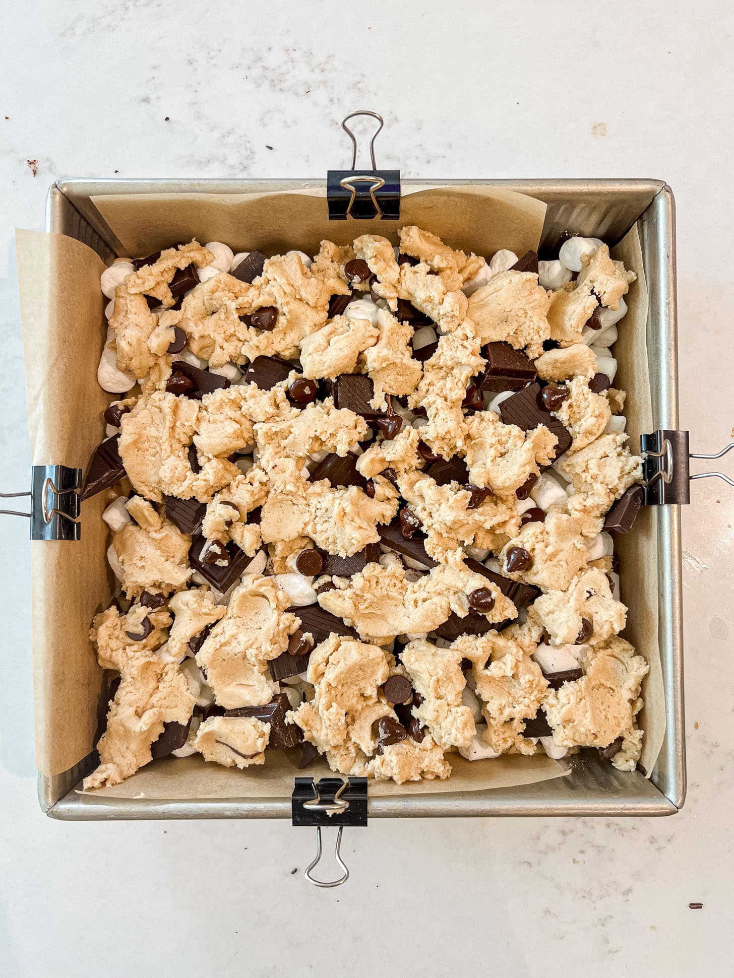 Spoonfuls of chocolate chip cookie dough dropped over the marshmallow and chocolate layer in a parchment-lined baking pan, ready to bake