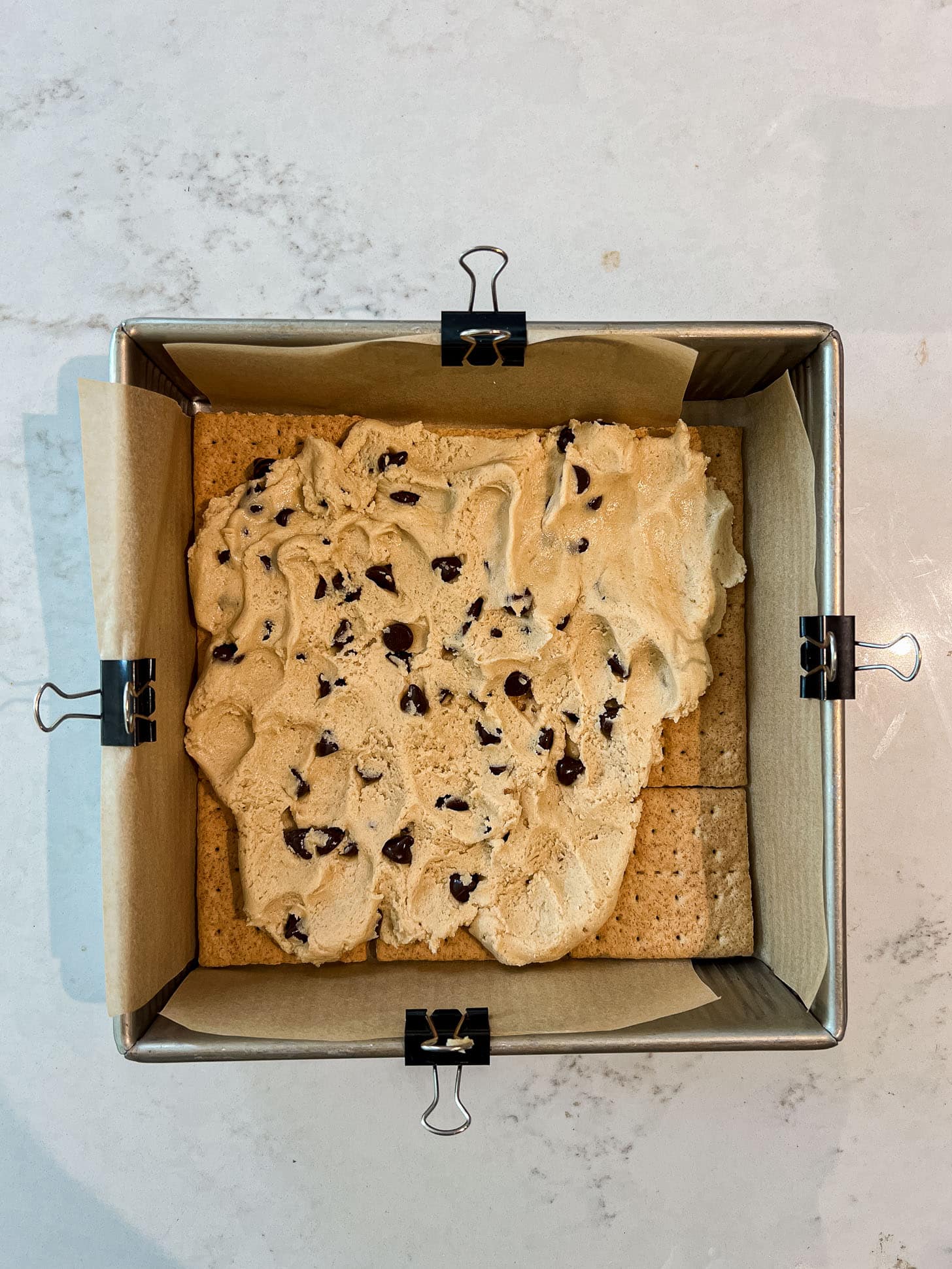 Chocolate chip cookie dough spread in an even layer over graham crackers in a parchment-lined square baking pan secured with binder clips