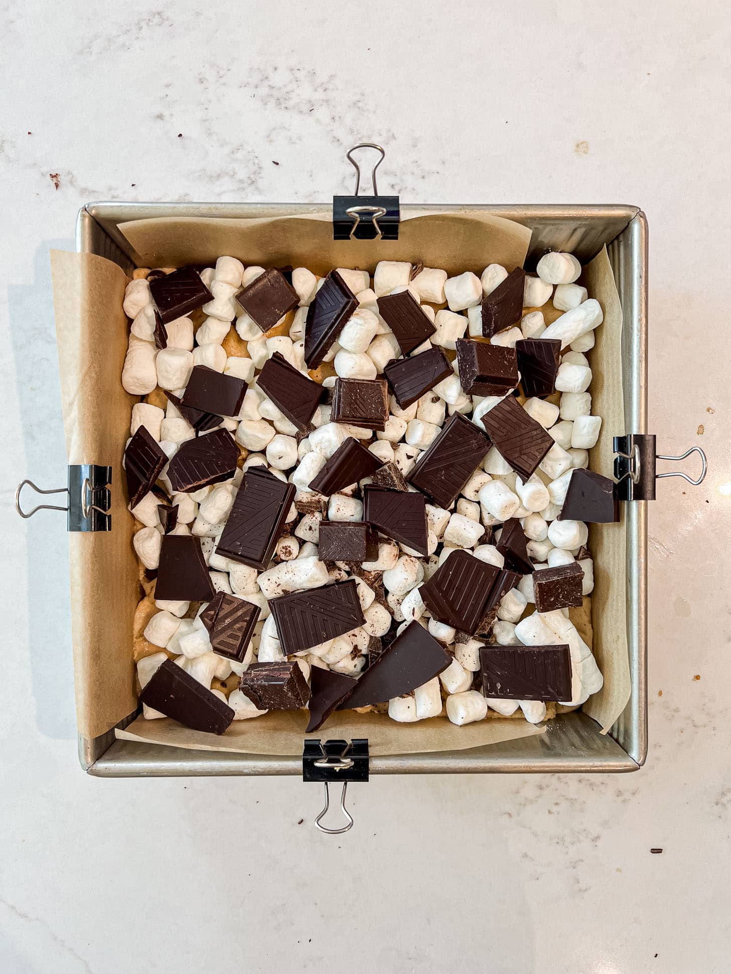 Mini marshmallows and broken chunks of dark chocolate scattered in an even layer over cookie dough in a parchment-lined baking pan