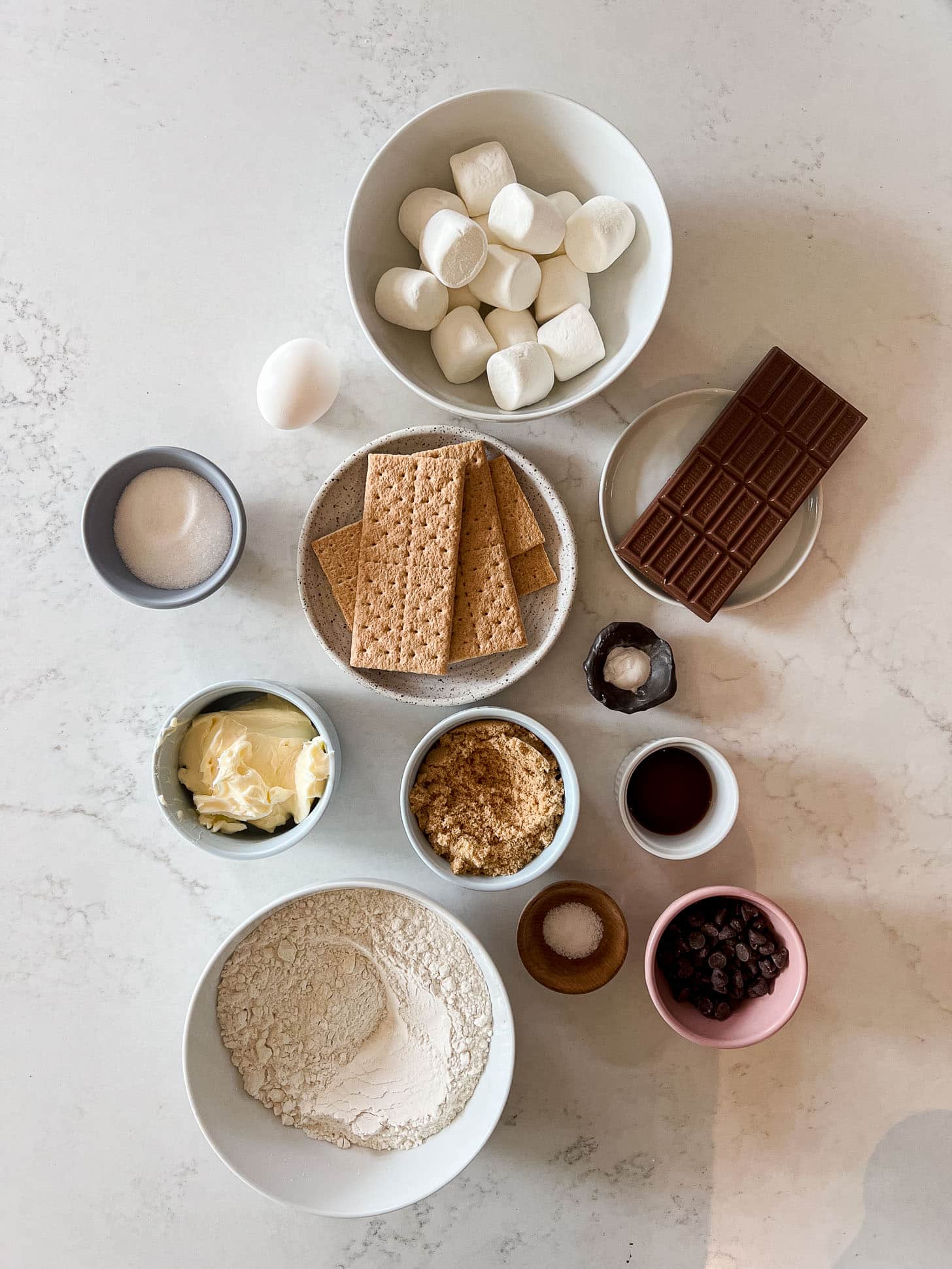 Flat lay of s'mores cookie bar ingredients including marshmallows, graham crackers, a chocolate bar, butter, brown sugar, flour, chocolate chips, an egg, vanilla extract, baking soda, and salt on a white marble surface