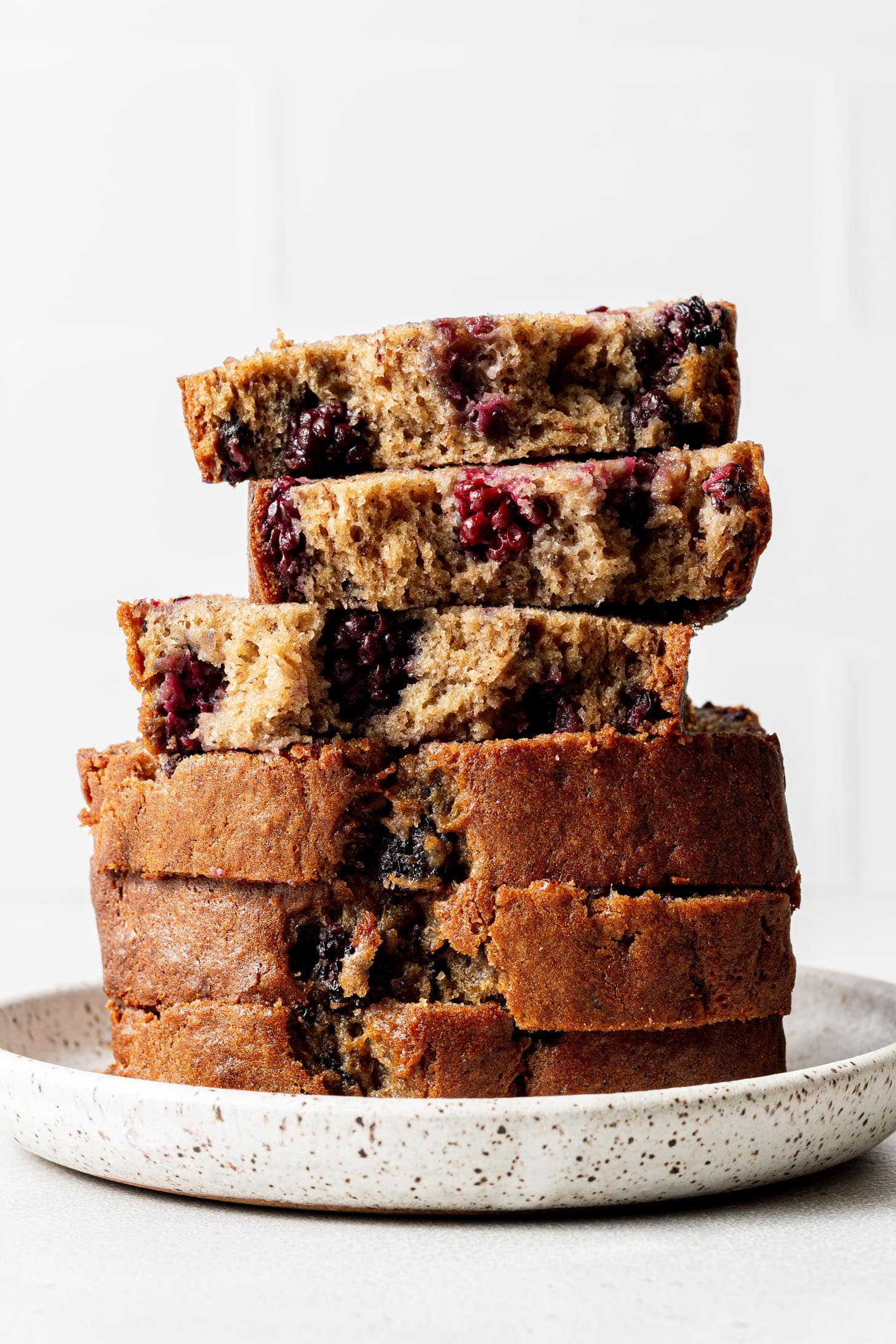 Four slices of blackberry banana bread stacked on a speckled white plate, showing the moist golden crumb packed with blackberries against a white background