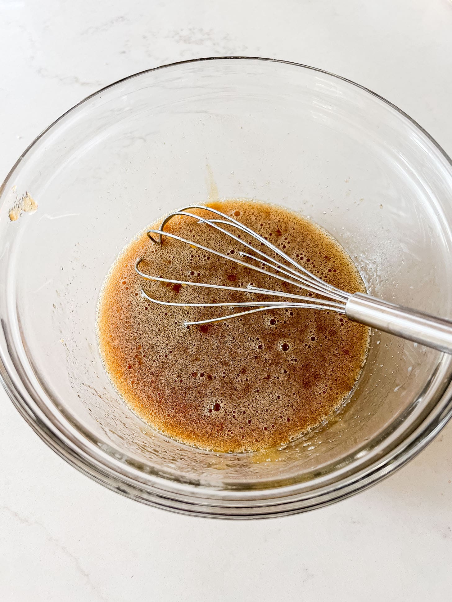Melted butter, eggs, and vanilla whisked together in a glass bowl on a white marble surface, forming the base of the banana bread batter