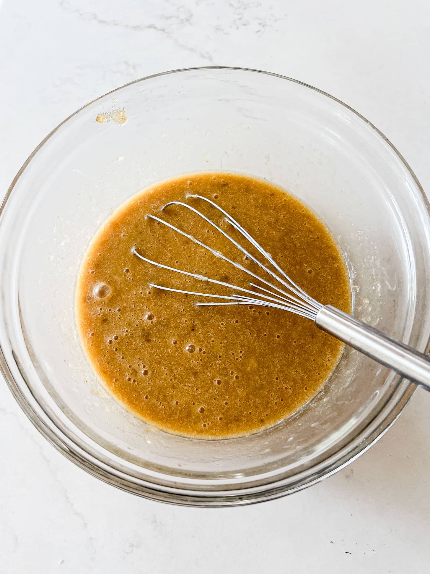 Banana bread wet ingredients including mashed banana and brown sugar whisked together in a glass bowl until smooth and golden brown