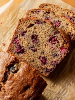 Close-up overhead view of sliced blackberry banana bread on parchment paper and a wooden board, showing the tender crumb loaded with blackberry pieces