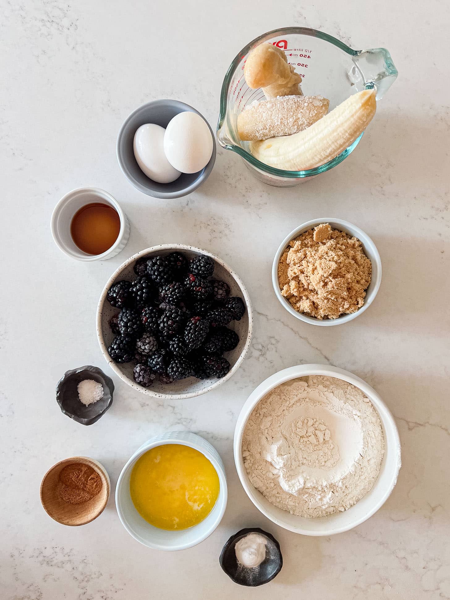 Flat lay of blackberry banana bread ingredients including fresh blackberries, ripe bananas, flour, brown sugar, eggs, melted butter, vanilla extract, cinnamon, nutmeg, salt, and baking soda on a white marble surface