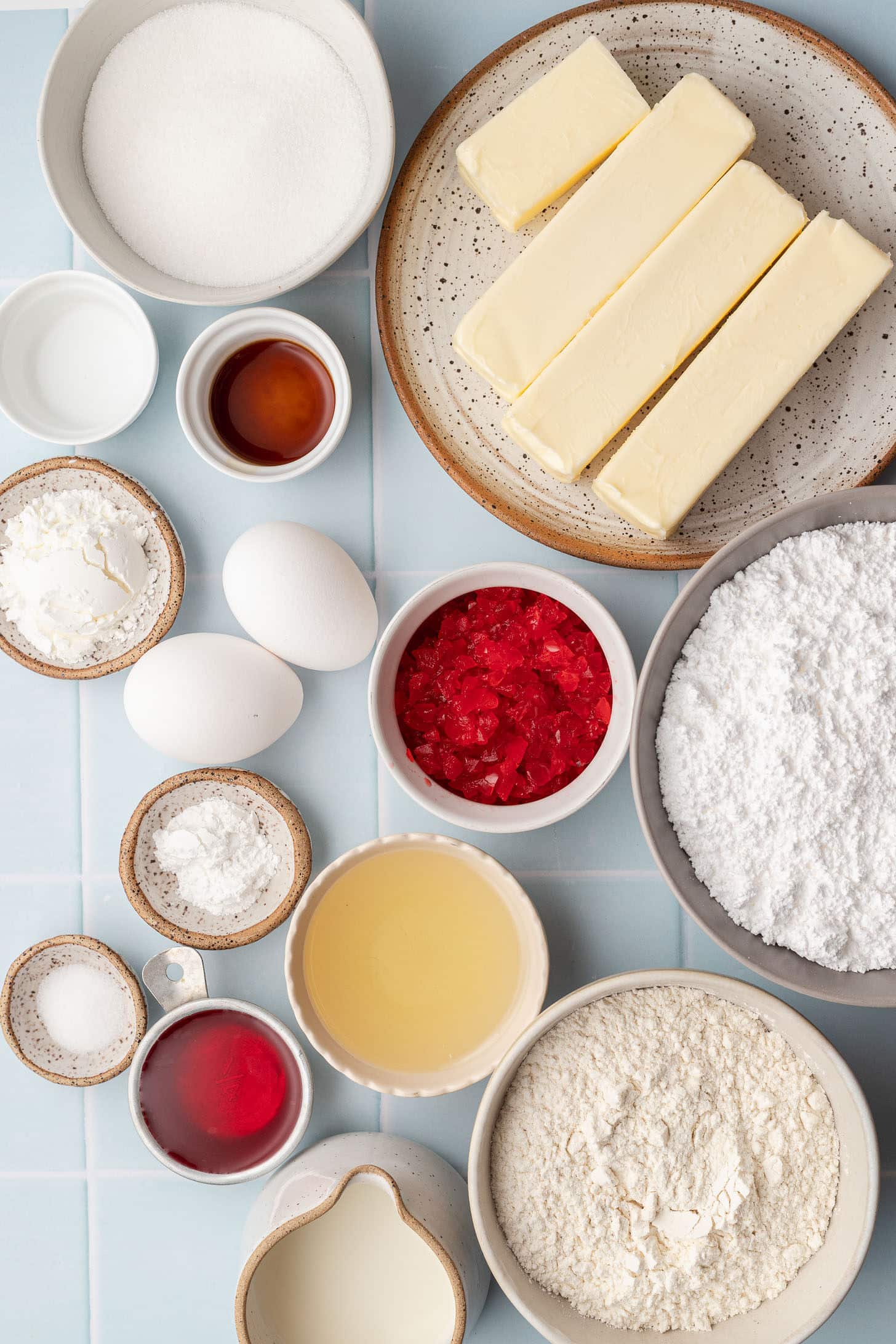Flat lay of cherry cupcake ingredients including butter, sugar, eggs, flour, cornstarch, baking powder, salt, diced maraschino cherries, maraschino cherry juice, milk, vanilla extract, and powdered sugar on a light blue tile surface