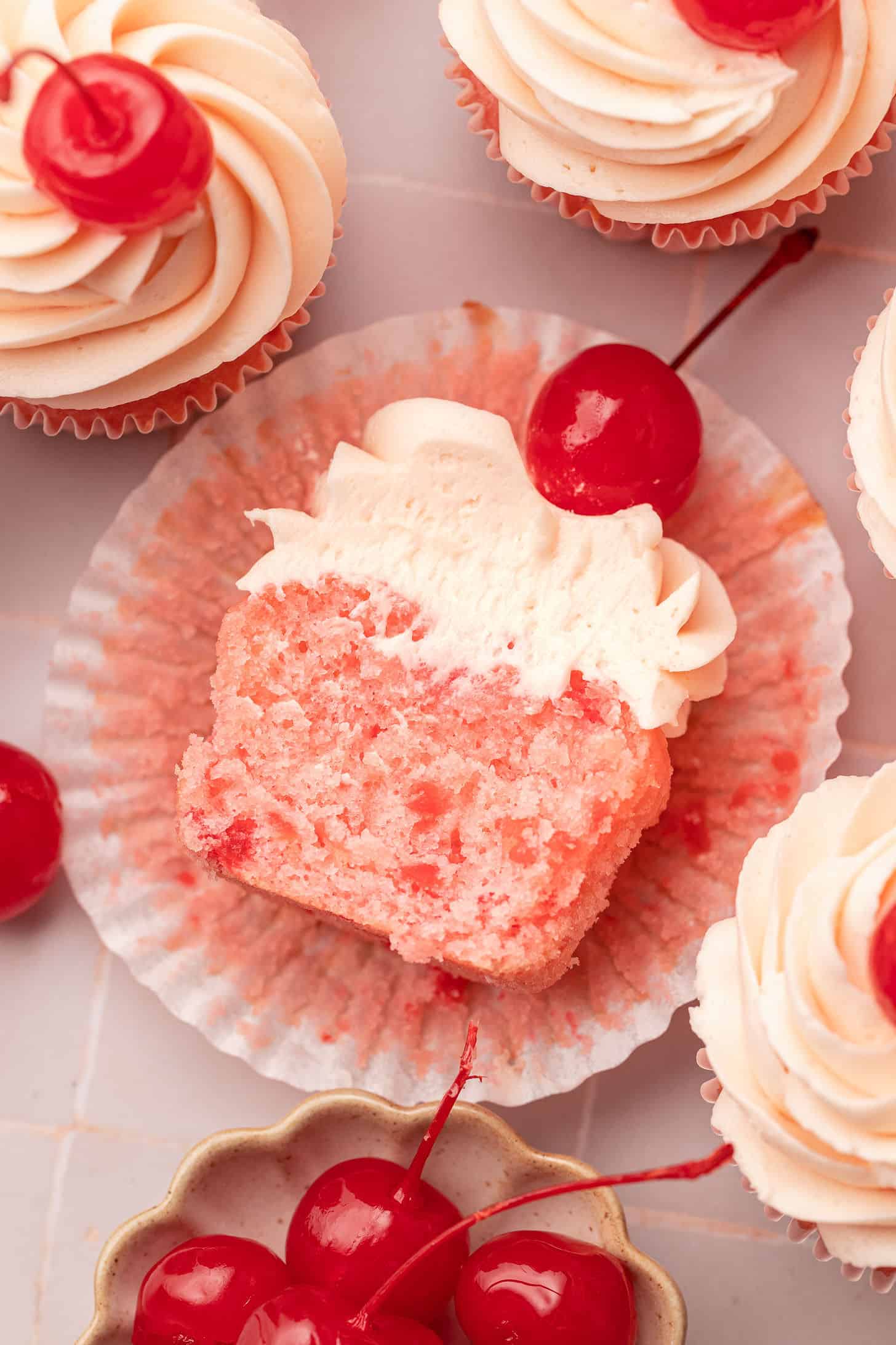 Overhead view of a split cherry cupcake showing the vivid pink interior with cherry pieces, surrounded by frosted cupcakes topped with maraschino cherries and a small bowl of maraschino cherries