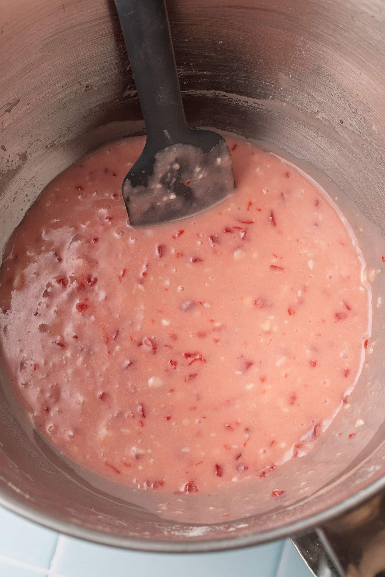 Smooth pink cherry cupcake batter with diced maraschino cherry pieces throughout, being folded with a black spatula in a stand mixer bowl