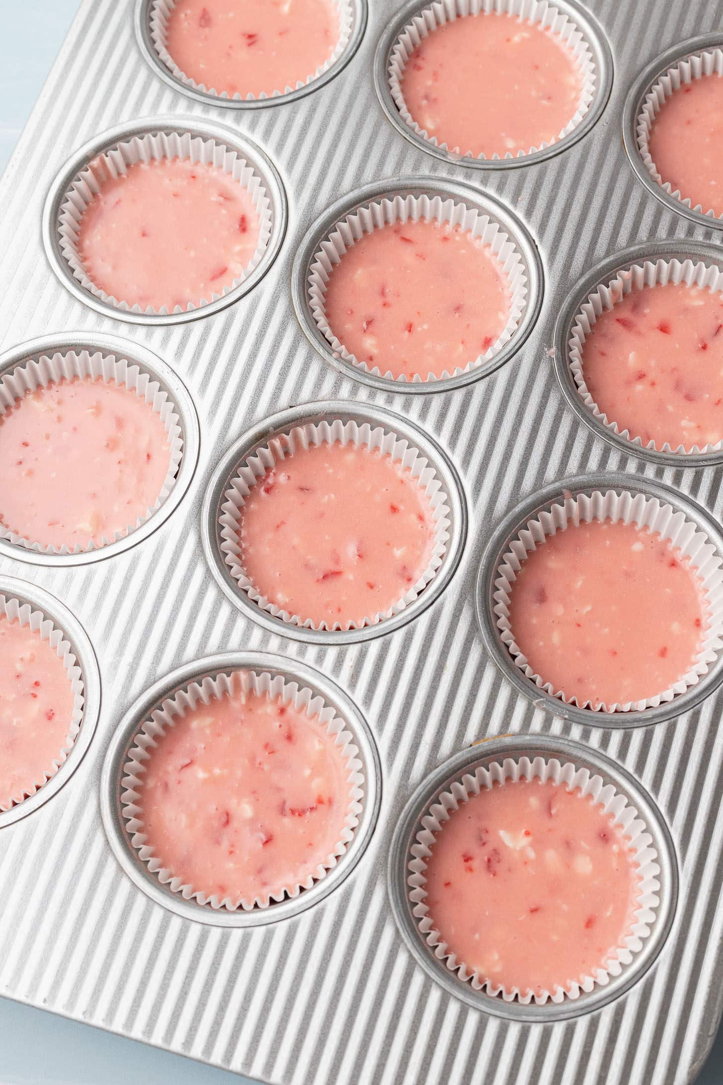 Pink cherry cupcake batter divided into a silver cupcake tin lined with white parchment liners, ready to bake