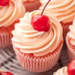 Close-up of cherry cupcakes with tall swirls of cherry almond buttercream and a glossy maraschino cherry on top, in pink cupcake liners on a wire cooling rack