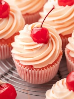 Close-up of cherry cupcakes with tall swirls of cherry almond buttercream and a glossy maraschino cherry on top, in pink cupcake liners on a wire cooling rack