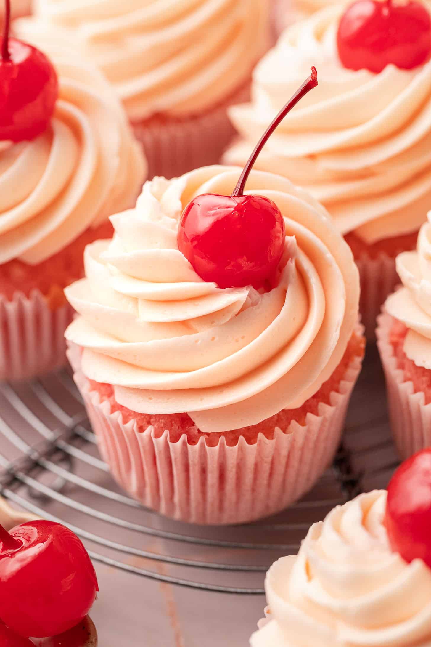 Close-up of cherry cupcakes with tall swirls of cherry almond buttercream and a glossy maraschino cherry on top, in pink cupcake liners on a wire cooling rack