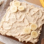 Overhead view of a banana cake spread with swirled cinnamon spiced buttercream and decorated with fresh banana slices, on parchment paper with a linen cloth and overripe banana in the background