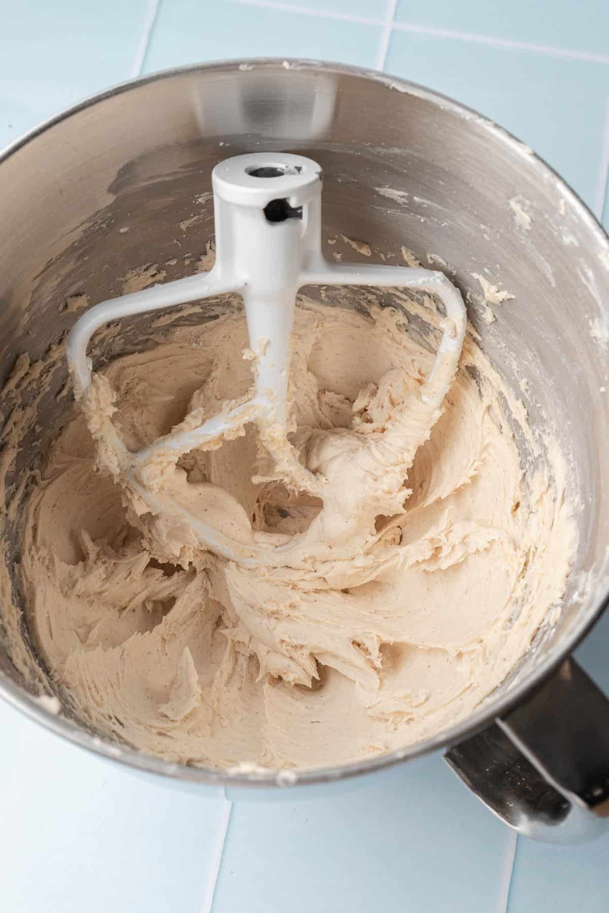 Fluffy cinnamon spiced buttercream whipped to a smooth, creamy warm beige color in a stand mixer bowl with the paddle attachment, on a light blue tile surface