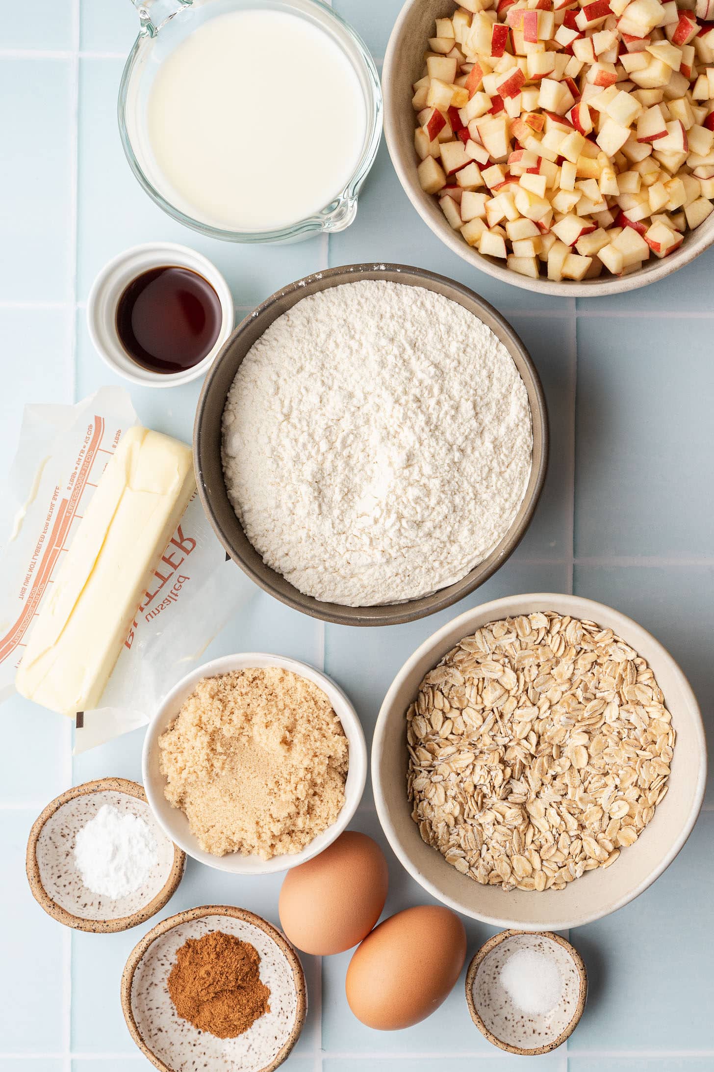 Flat lay of apple oat muffin ingredients including diced apples, rolled oats, flour, brown sugar, butter, eggs, milk, vanilla extract, cinnamon, baking powder, and salt on a light blue tile surface