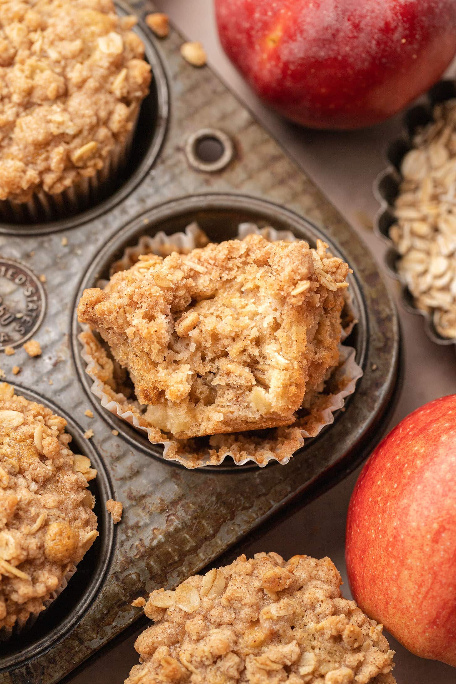 An apple oat streusel muffin broken in half and resting in a vintage muffin tin, showing the soft apple-studded crumb inside, with red apples and rolled oats nearby