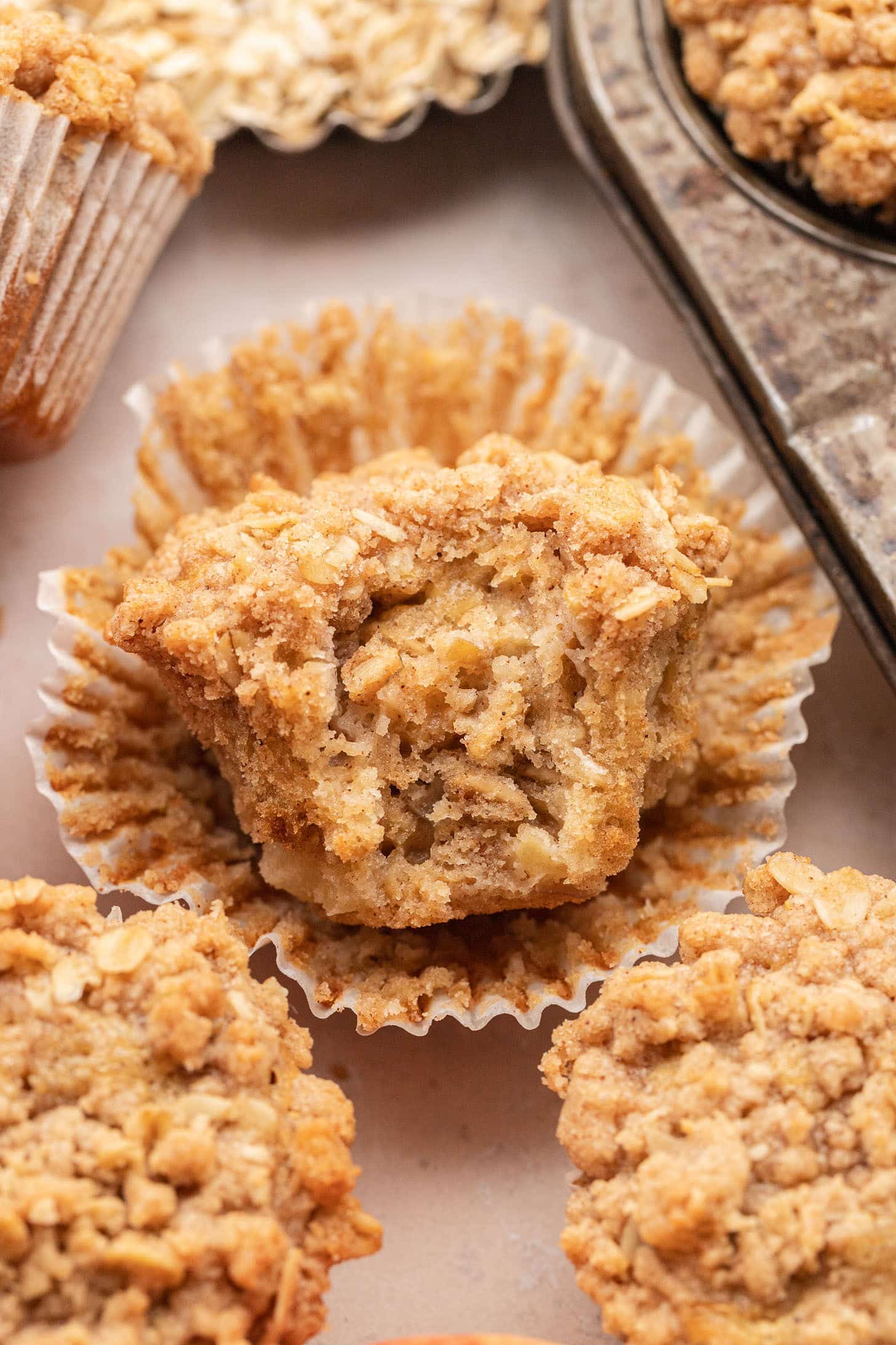 An apple oat muffin with its wrapper peeled back to reveal the soft, moist interior with apple pieces and oats, surrounded by more streusel-topped muffins and a vintage muffin tin